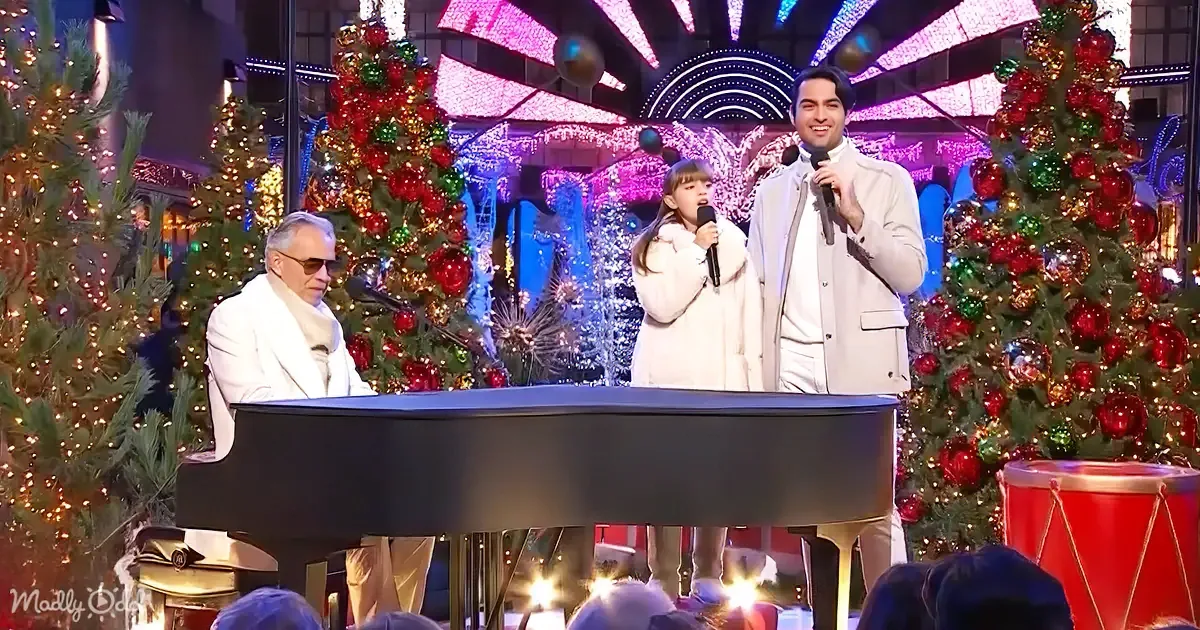 A man in white plays piano while a young girl and another man sing in front of festive Christmas trees.