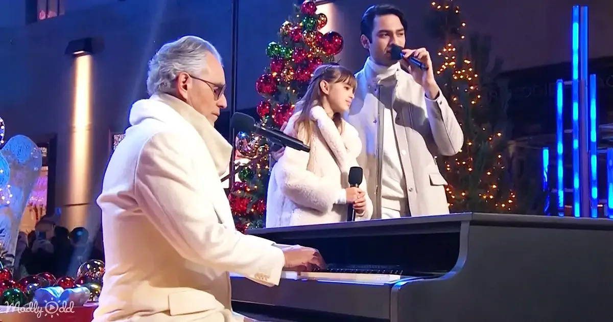 A man in white plays piano while a young girl and another man sing in front of a Christmas tree.