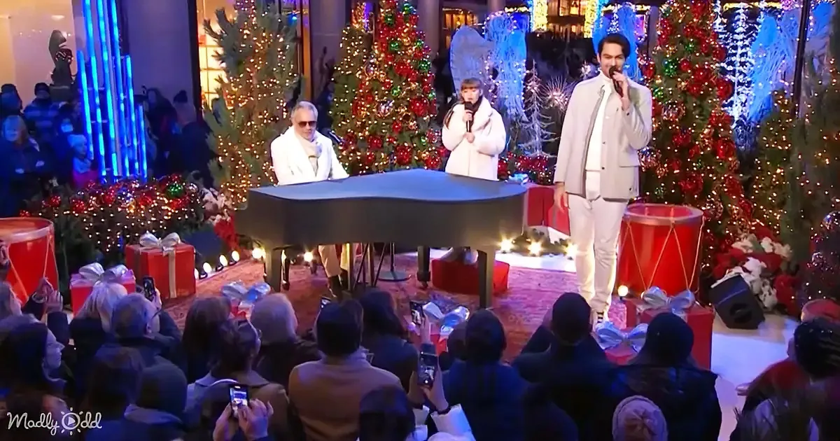 A man in white sings into a microphone on stage with a piano and Christmas decorations.