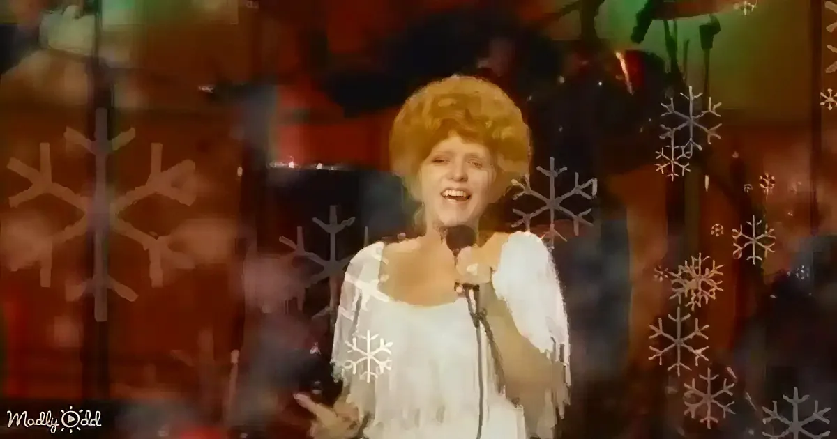 A person in a white outfit with a large yellow hat and wings stands in front of a microphone, smiling.
