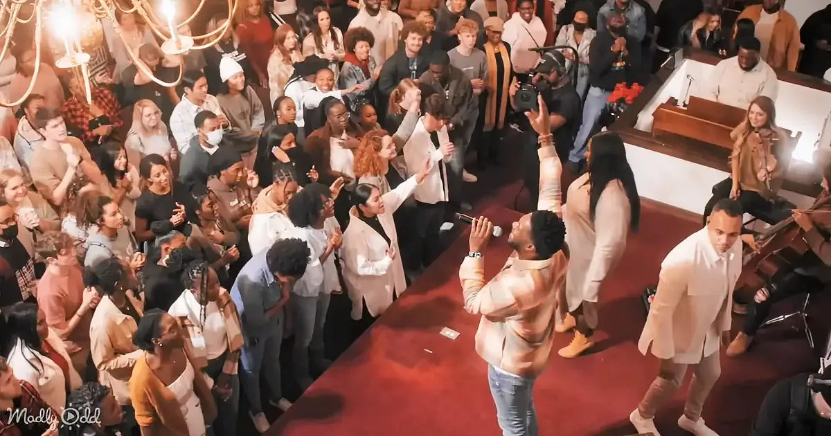 A man in a church holds a microphone, singing to a crowd of people, some clapping and smiling.