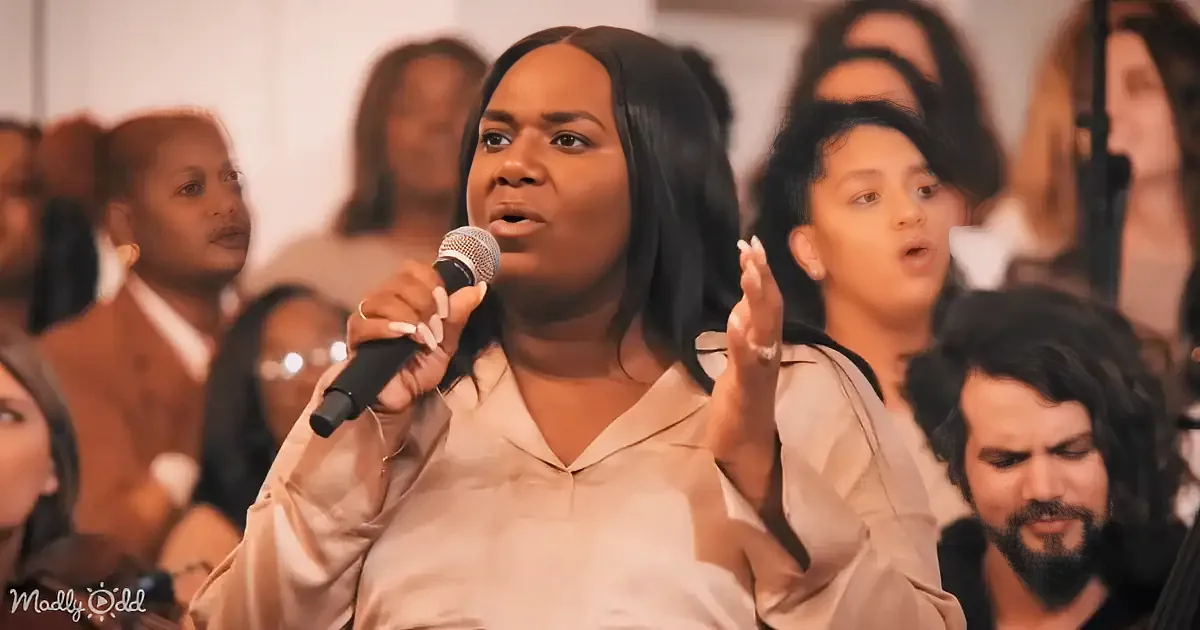 A woman in a beige shirt sings into a microphone, surrounded by a choir.