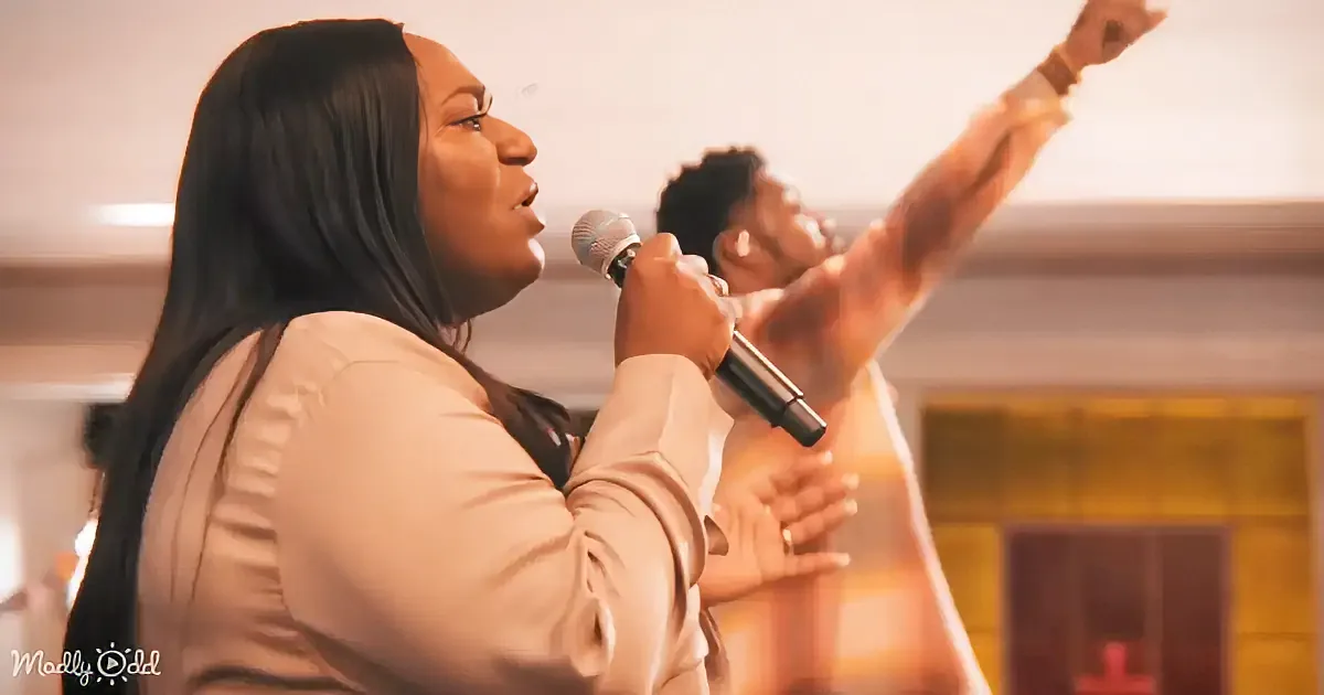 A woman sings passionately into a microphone, raising her arm in the air, with another person behind her.