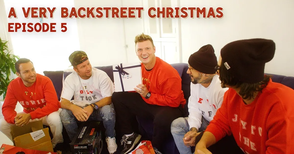 A man in a red shirt holds a gift box, smiling, surrounded by friends in casual attire.