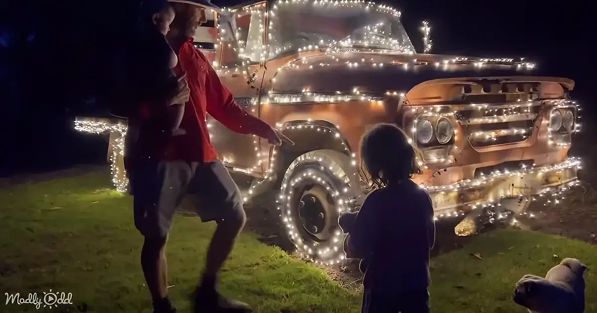 A man and child admire a vintage truck adorned with string lights at night.