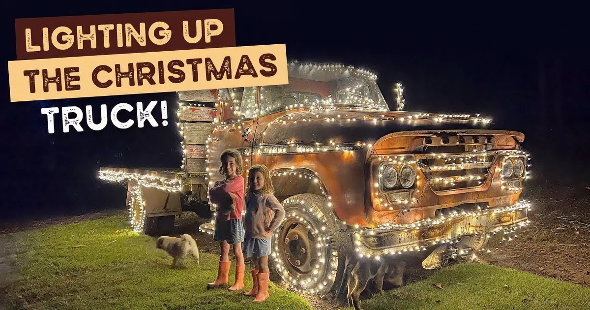 An old truck adorned with lights sits on a grassy field at night, with two children and a dog nearby.