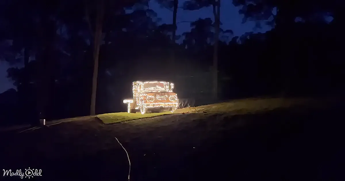 A vintage truck is adorned with festive lights, glowing brightly against a dark, tree-lined background.