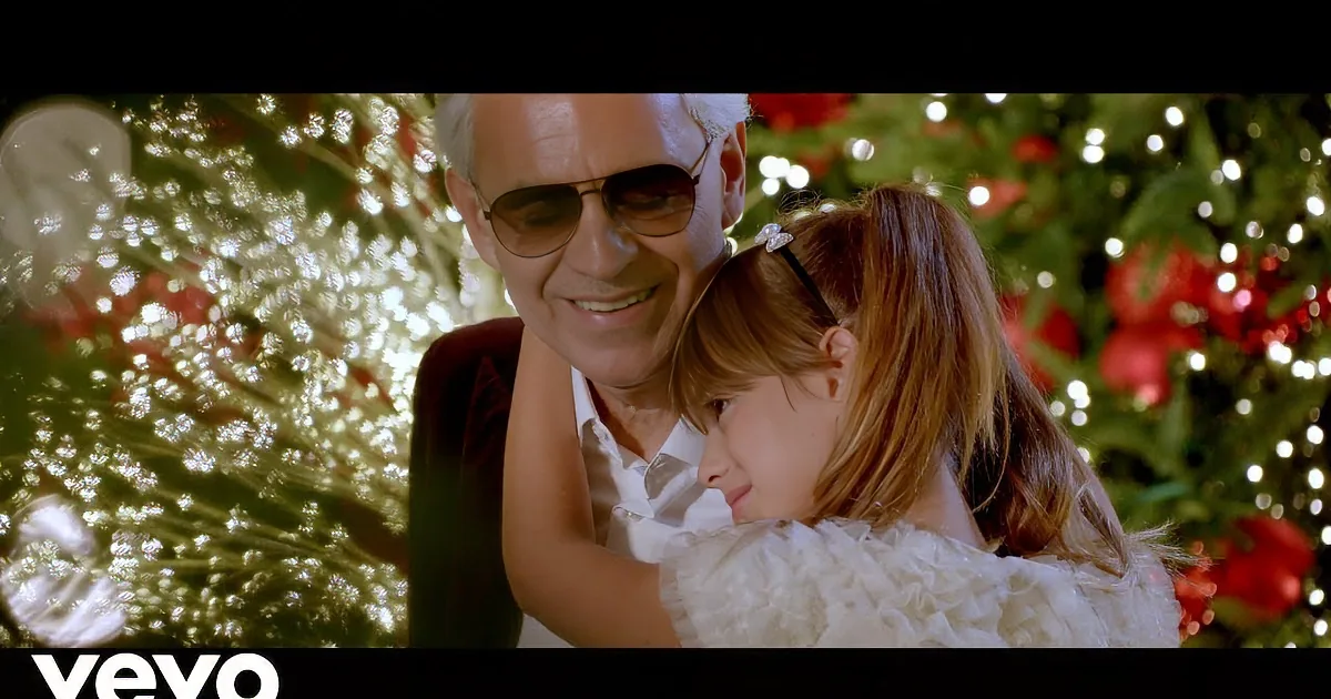 A man in a suit and sunglasses smiles at a young girl in a white dress, both in front of a festive Christmas tree.