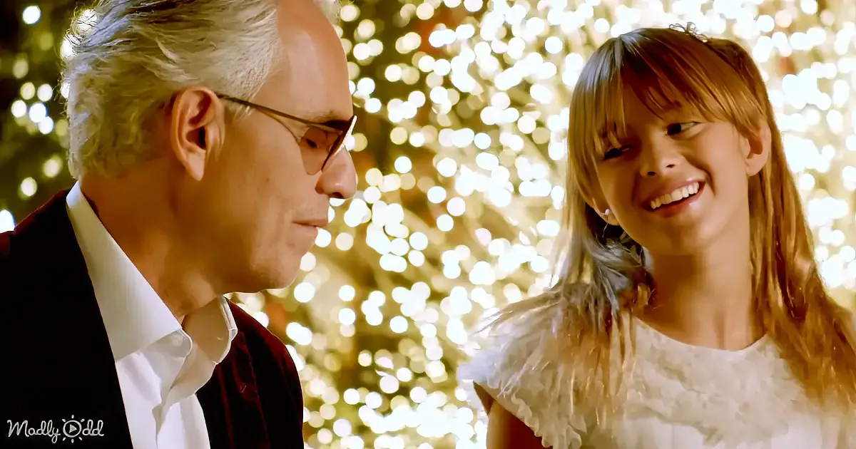 A man in a suit and a young girl in a white dress smile at each other against a backdrop of twinkling lights.