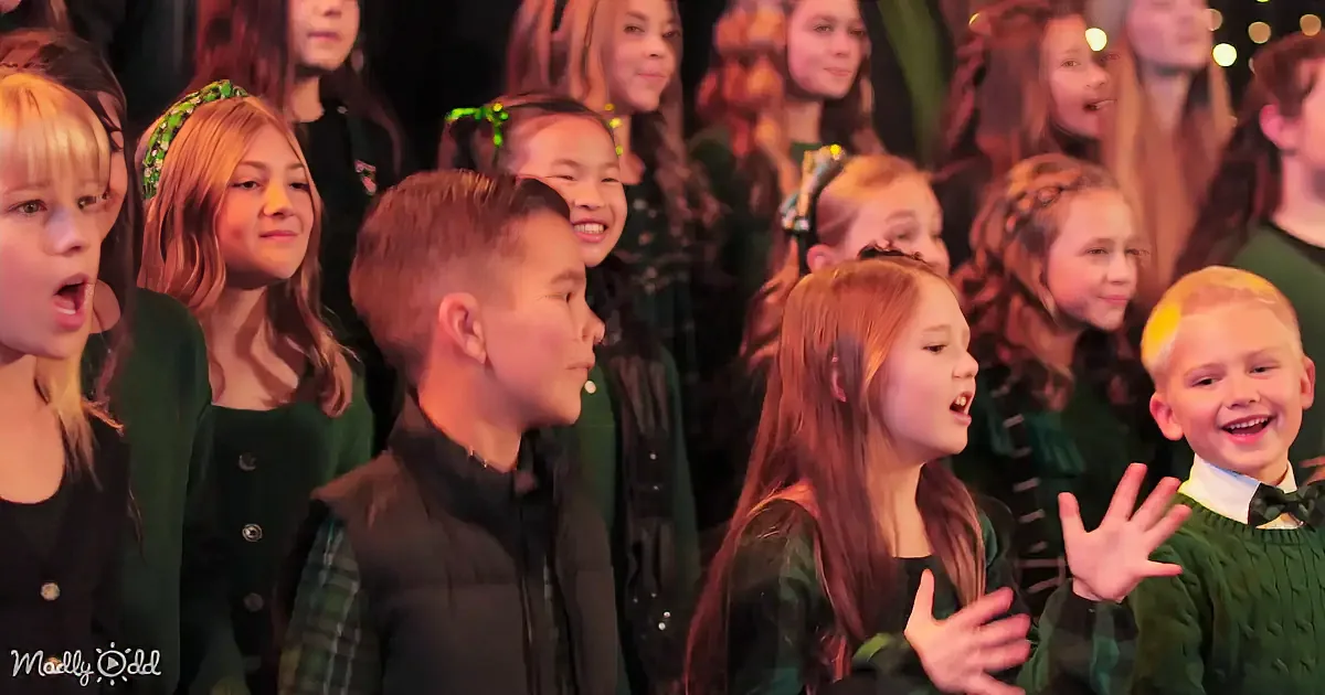 A young boy in a black jacket stands among a group of children in green, singing and smiling.