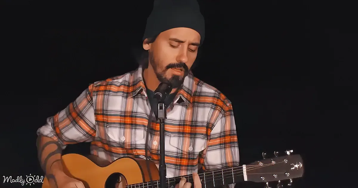 A man in a plaid shirt and beanie plays an acoustic guitar while singing into a microphone.