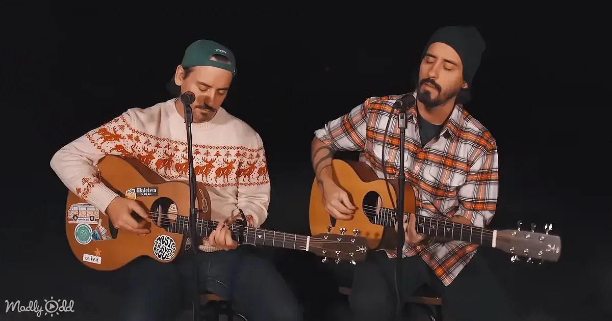Two men play acoustic guitars, one in a sweater with reindeer, the other in a plaid shirt, both singing into microphones.
