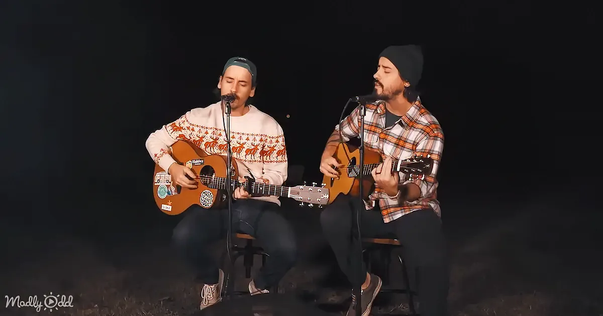 Two men play acoustic guitars and sing into microphones, wearing winter clothing, against a dark background.