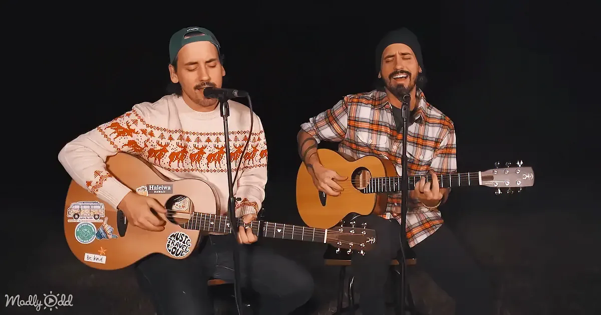 Two musicians play acoustic guitars and sing into microphones, one wearing a reindeer sweater, the other a plaid shirt.