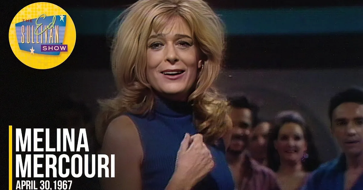 A woman with blonde, shoulder-length hair and a blue turtleneck smiles while holding her hand near her chest.