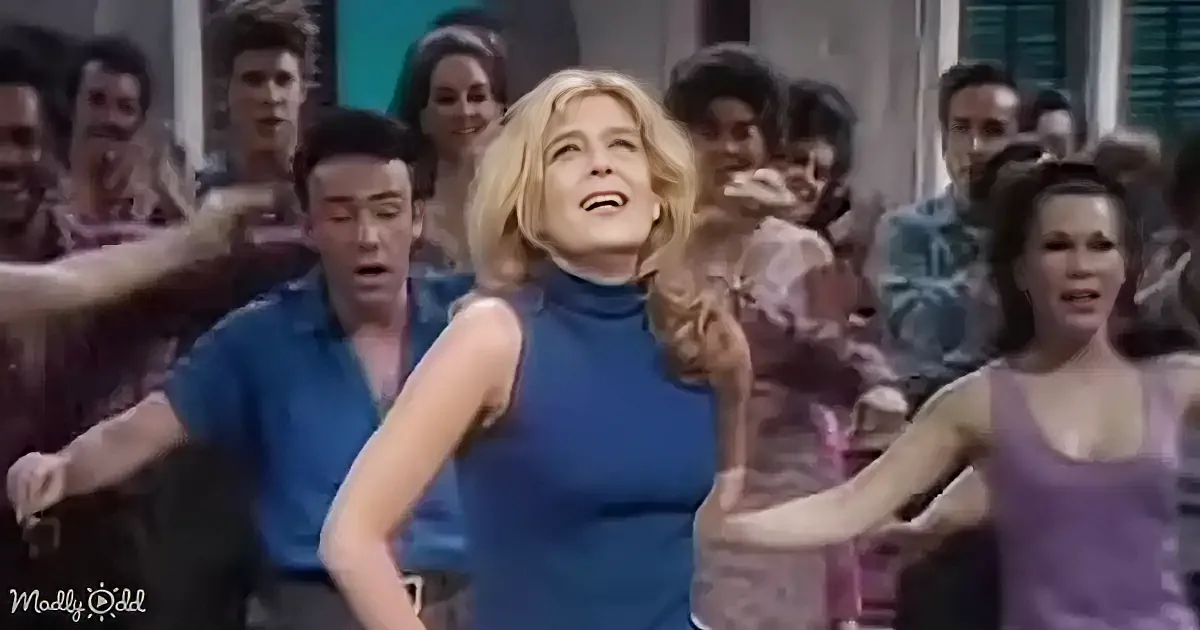 A woman in a blue dress dances joyfully in front of a crowd, surrounded by people in colorful outfits.