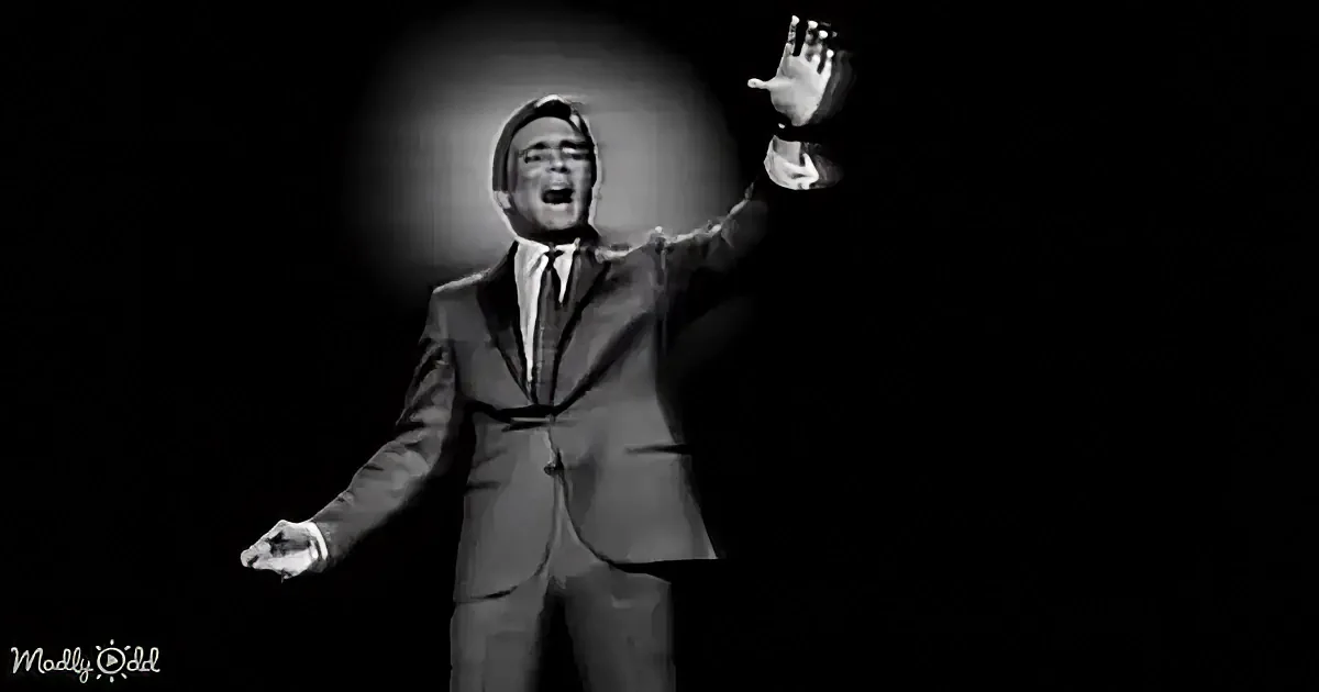 A man in a suit and tie is singing passionately on stage with his arms outstretched.