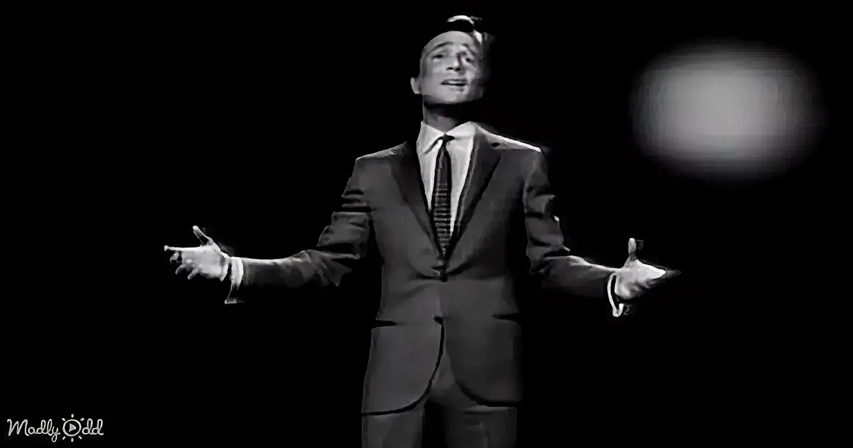 A man in a suit and tie stands with arms outstretched, singing on stage.