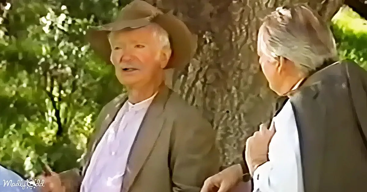 An older man in a hat and suit stands next to another older person in a white shirt and glasses, both outdoors near a tree.