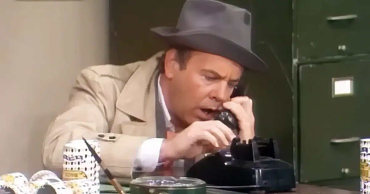 A man in a trench coat and fedora is talking on a phone while holding a pen and looking at a file cabinet.