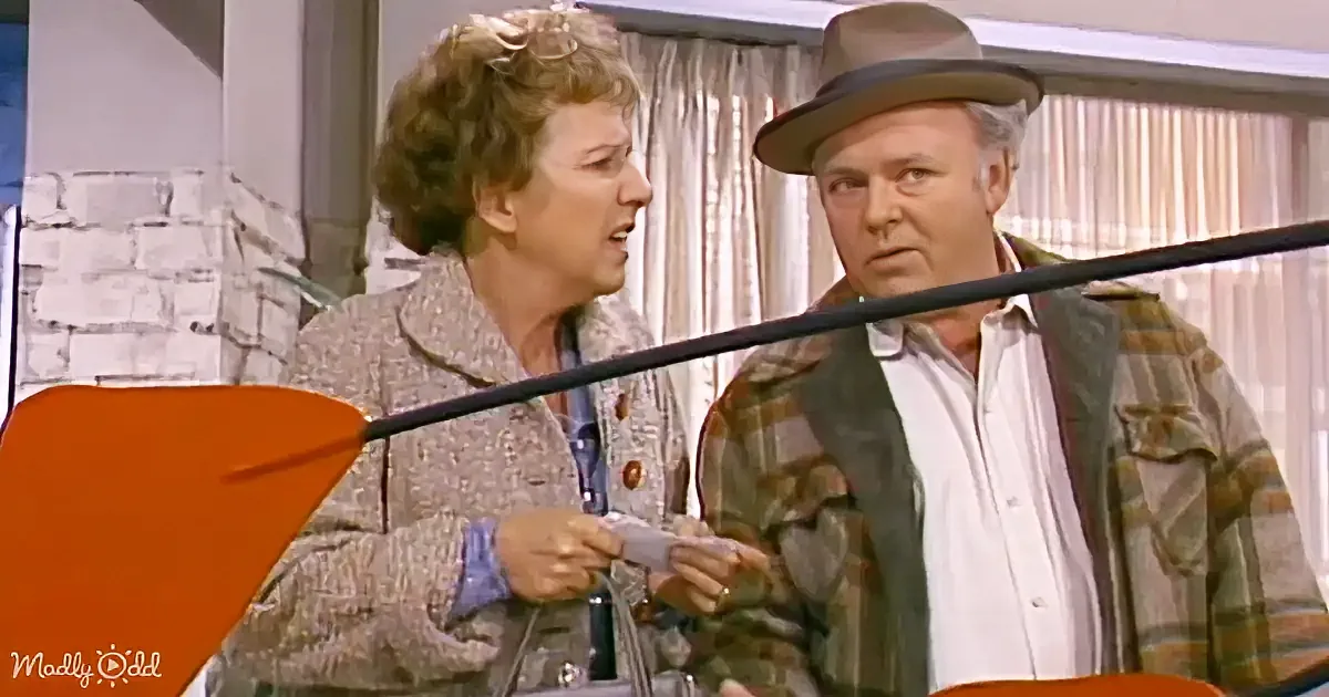 A woman in a patterned jacket holds a card, talking to a man in a hat and plaid shirt.