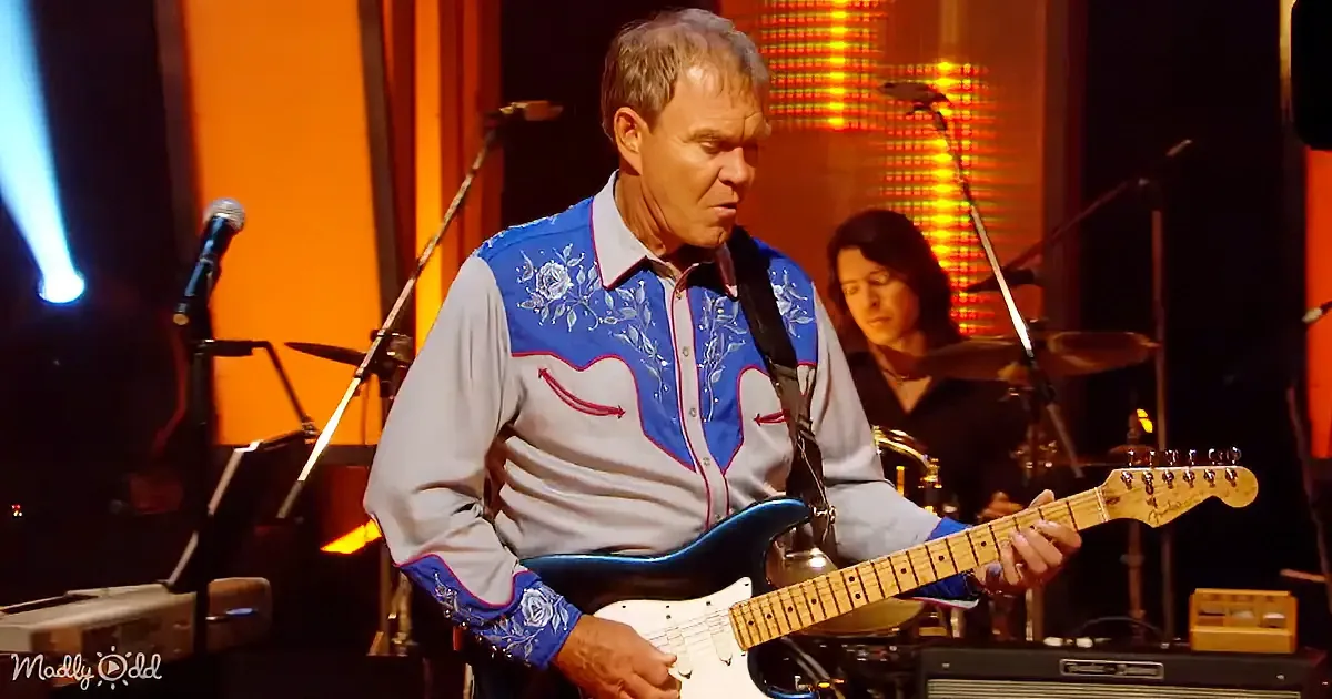 A man in a blue and white embroidered shirt plays a white electric guitar on stage.