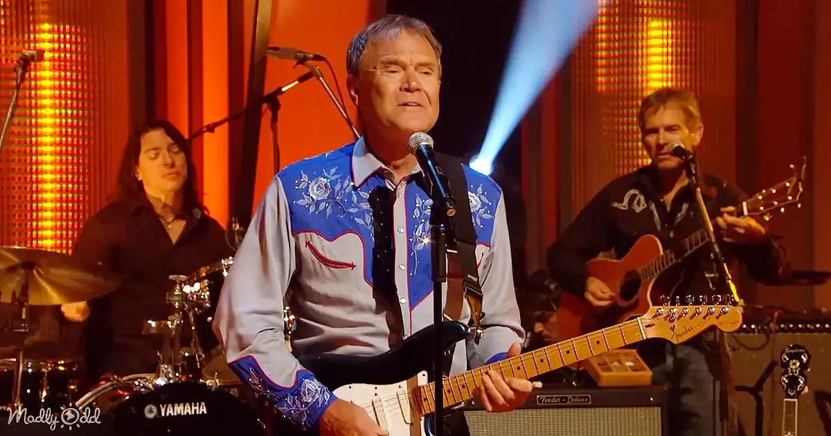 A man in a blue and white embroidered shirt plays guitar and sings into a microphone on stage.