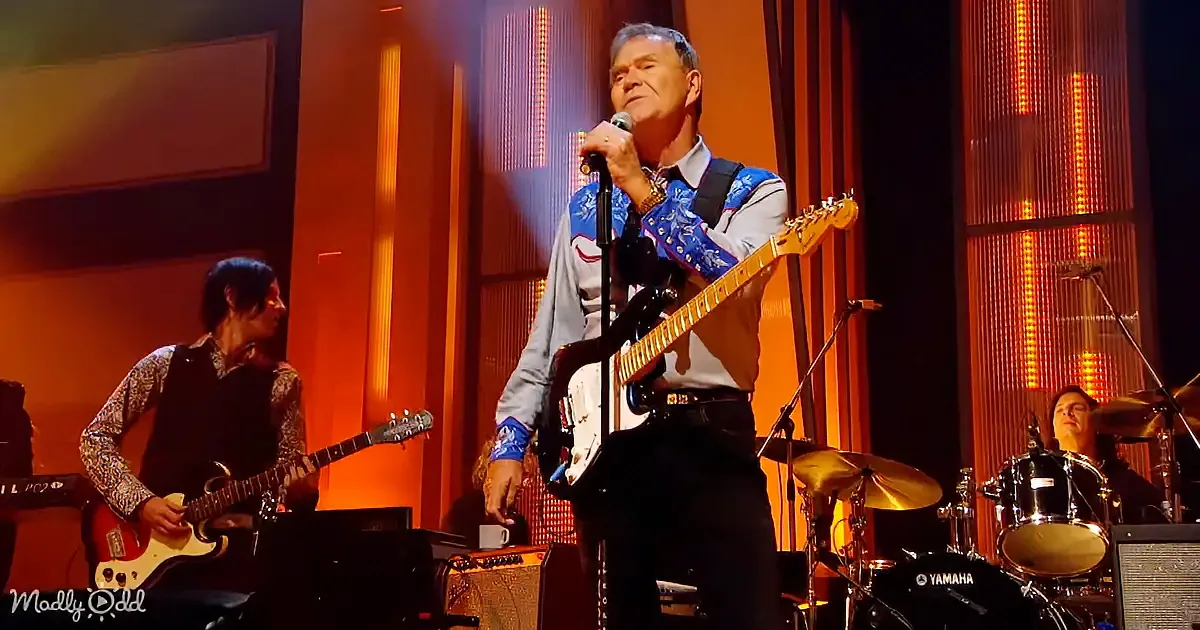 A man in a blue and white shirt sings into a microphone while playing a guitar on stage.