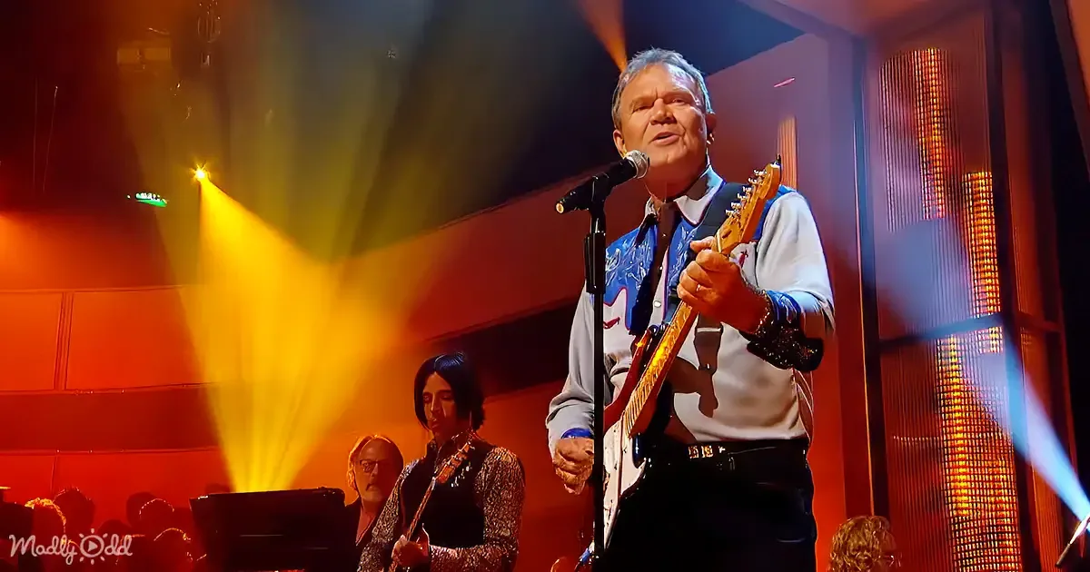 A man in a white shirt and blue vest plays guitar and sings on stage with a band.