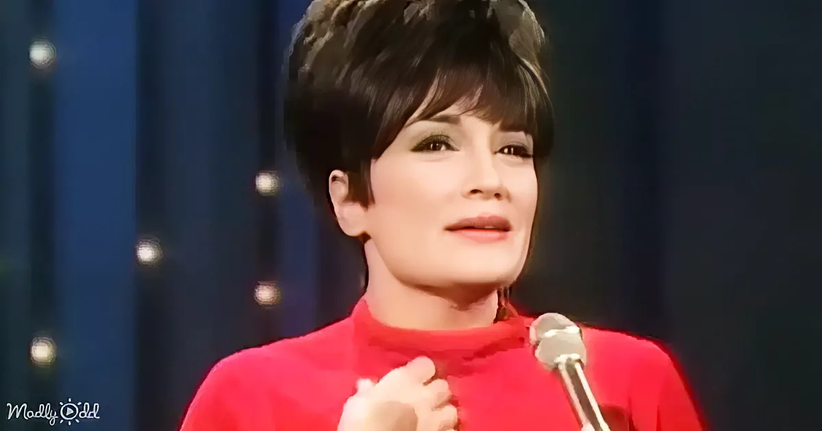 A woman in a red top speaks into a microphone, with a dark background and small lights.