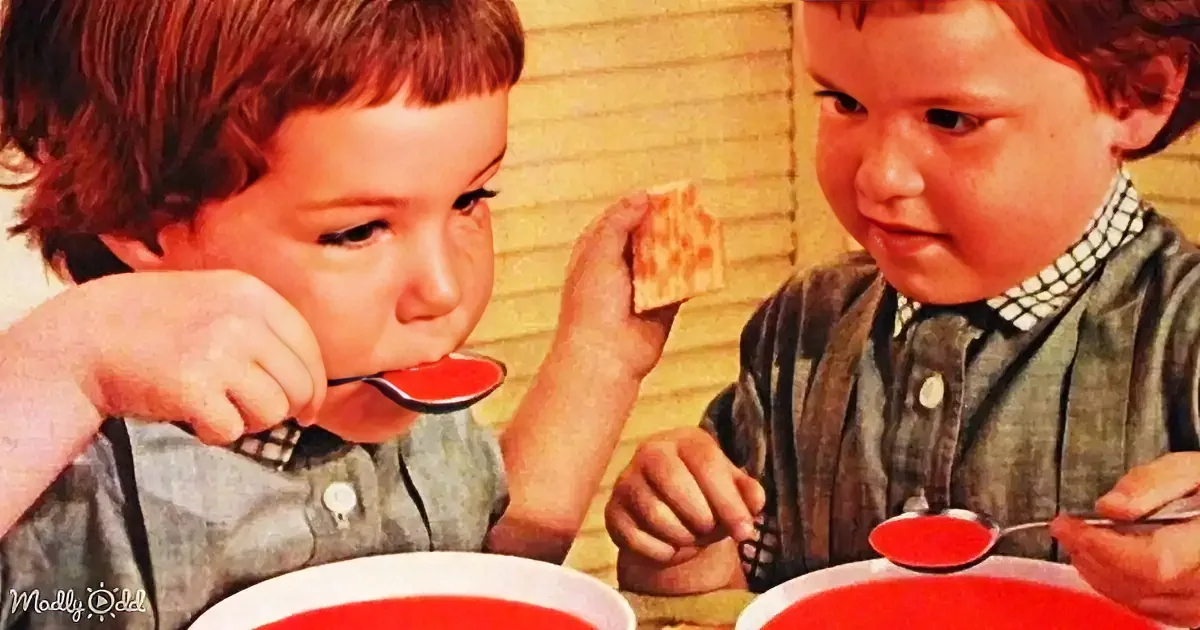 Two young boys are eating soup and crackers, one holding a spoon and the other a cracker.