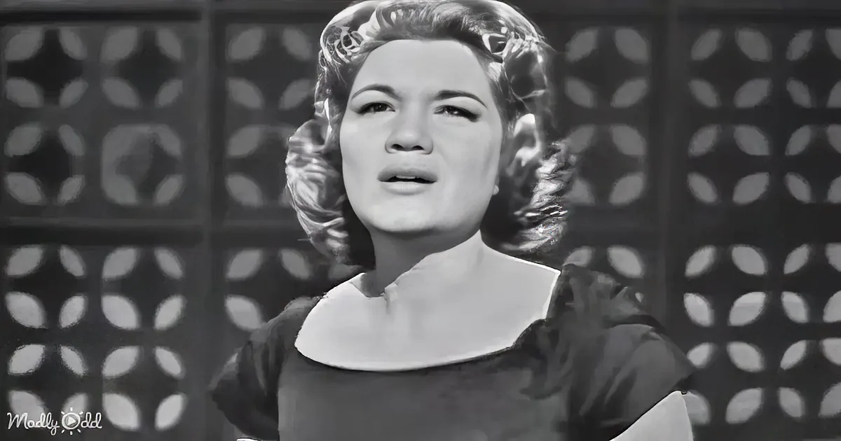 A woman with curly hair in a black dress stands in front of a patterned background, mouth slightly open.