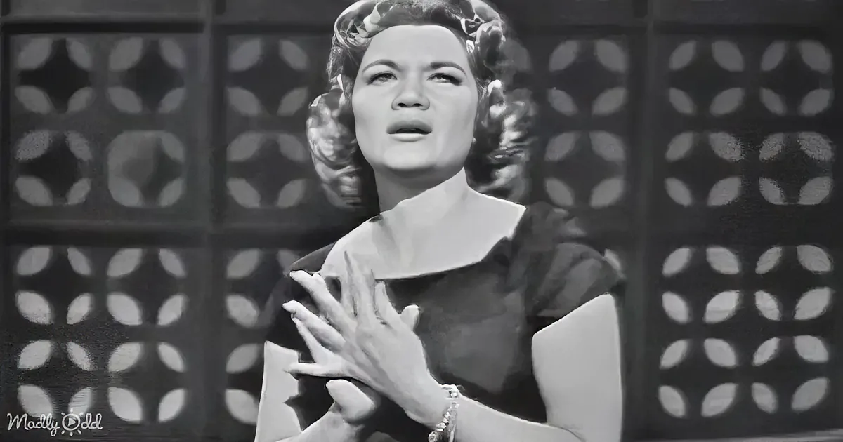 A woman with voluminous hair and a dark dress is singing passionately, hands clasped near her chest.