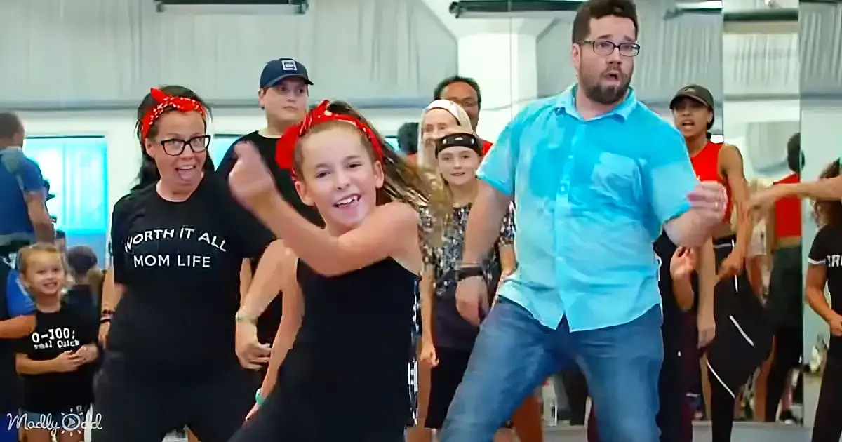 A young girl in a black outfit dances enthusiastically with a man in a blue shirt, surrounded by other children and...