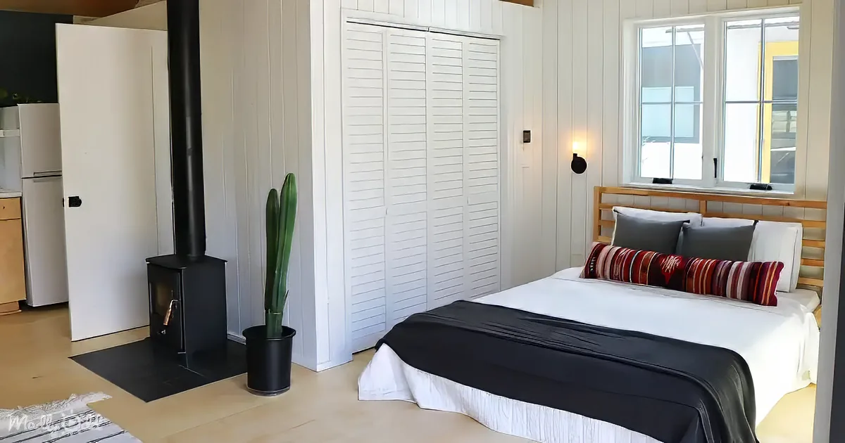 A cozy bedroom with a bed, white closet, wood stove, and large window.