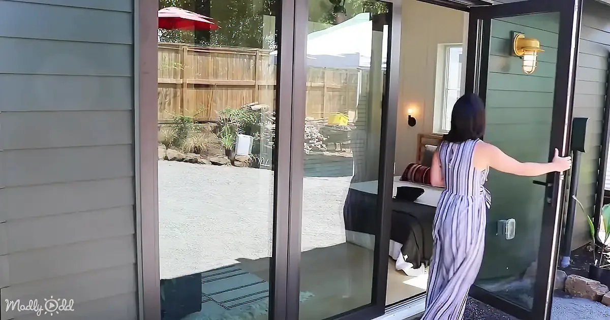 A woman in a striped dress opens a glass door to a backyard with a wooden fence and patio.
