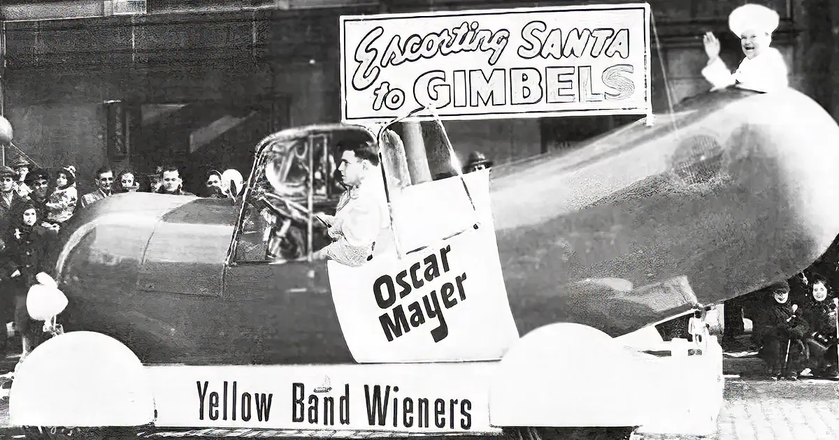 A man in a chef's hat drives a hot dog-shaped car in a parade, promoting Oscar Mayer Yellow Band Wieners.