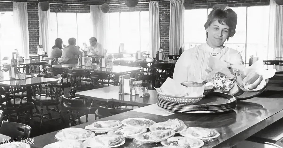 A woman in a white apron holds a basket of food at a restaurant counter with plates of food in front.