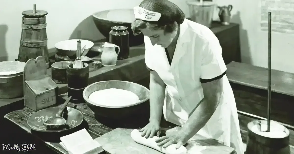 A chef in a white uniform kneads dough on a wooden surface in a kitchen setting.