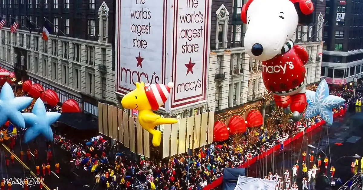 A large yellow balloon resembling a cartoon character floats above a festive parade with a Snoopy balloon and a crowd of...