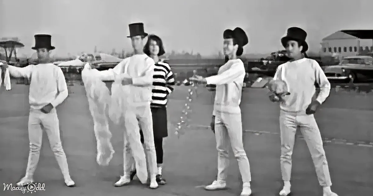 A group of five people in white outfits and hats stands in a parking lot, with a woman in a striped shirt in the center.
