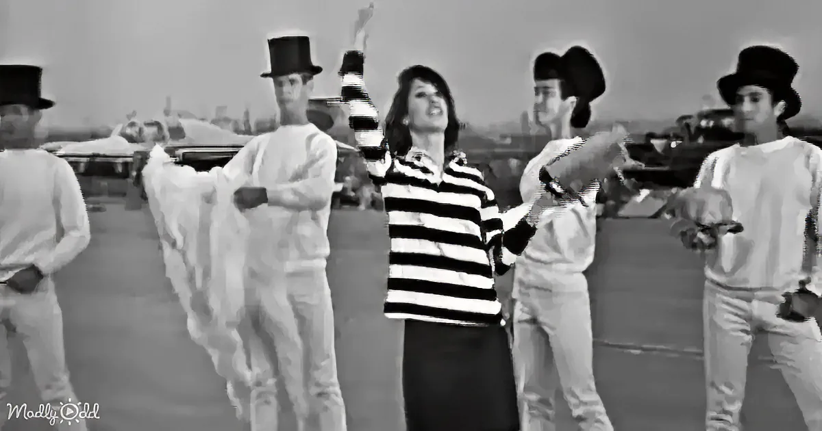 A woman in a striped sweater dances with a group of men in white suits and top hats on an airport tarmac.