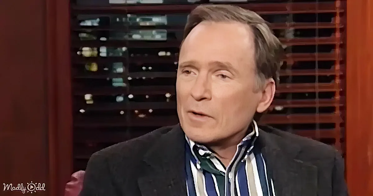 A man in a suit and striped shirt is speaking, with a wine rack and wooden blinds in the background.