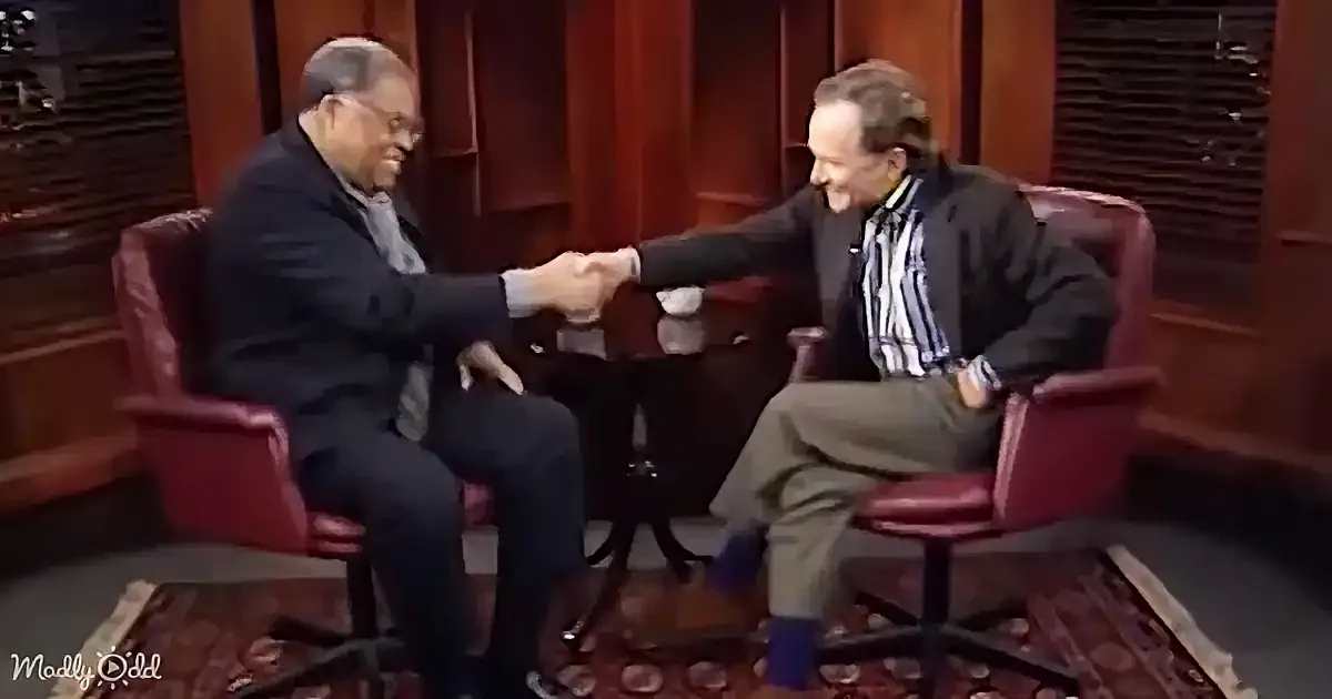 Two men in suits shake hands in a studio setting, both smiling.
