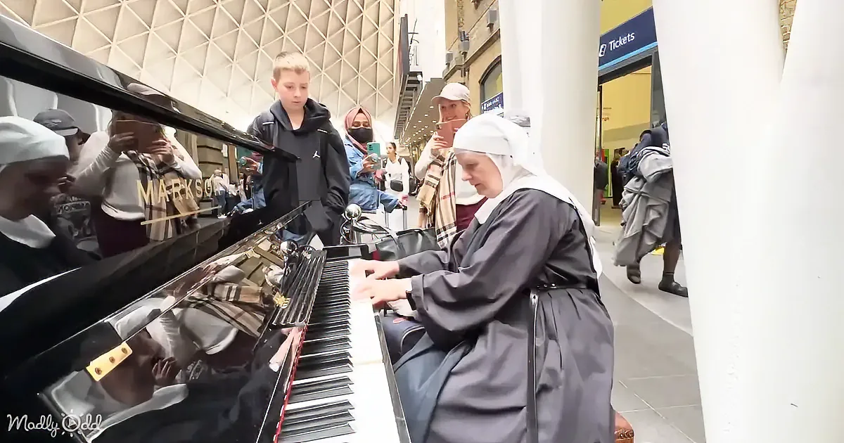 A nun plays a piano in a busy train station, surrounded by onlookers taking photos.