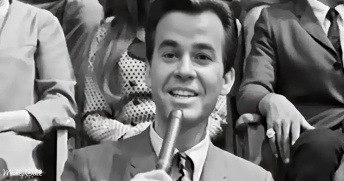 A man in a suit holds a cigar, smiling, with others in the background.