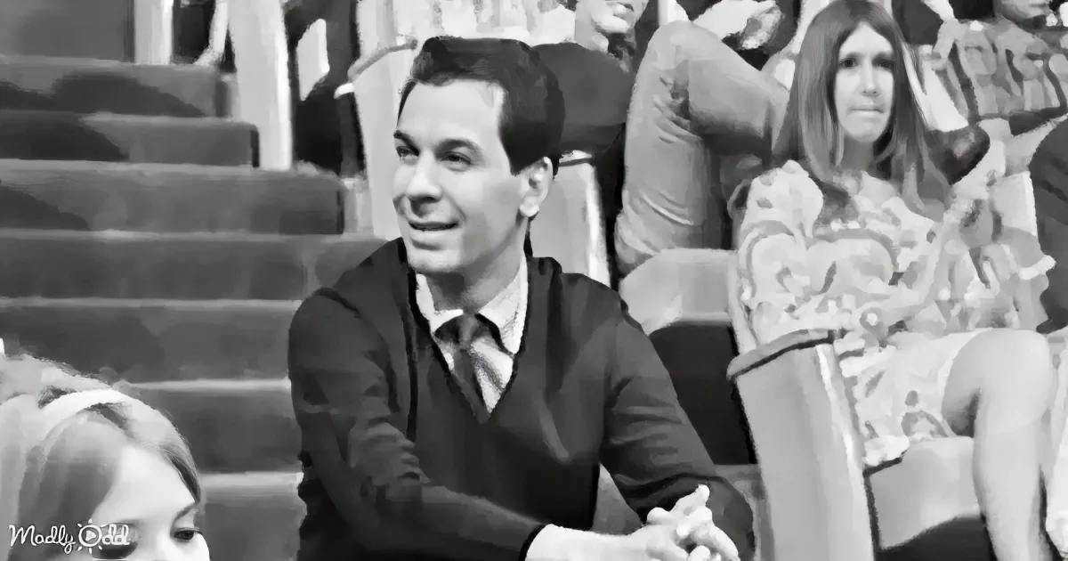 A man in a sweater and tie smiles while sitting in a stadium-like setting with other people around him.