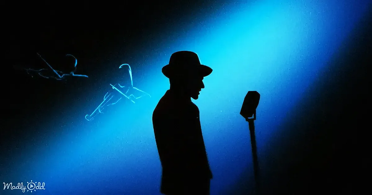 A silhouette of a man in a hat stands beside a microphone under a blue spotlight.