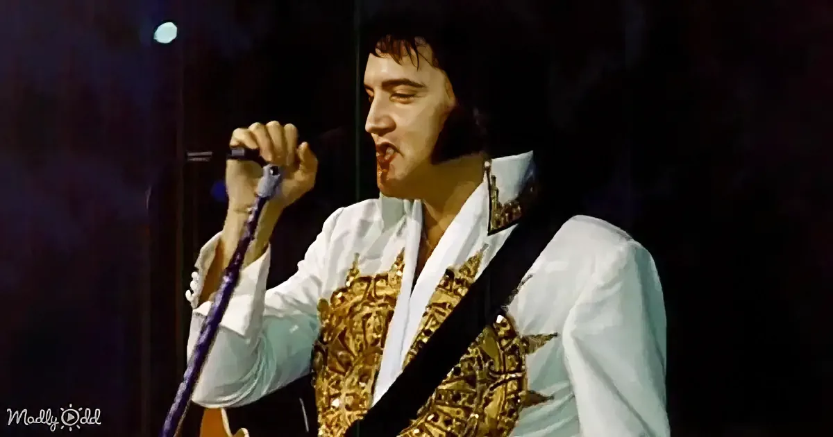 Elvis Presley in a white shirt and gold vest, holding a microphone and a guitar, performing on stage.