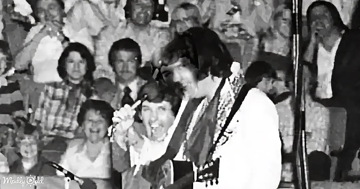 A man with long hair and a guitar smiles on stage, surrounded by a crowd of people.